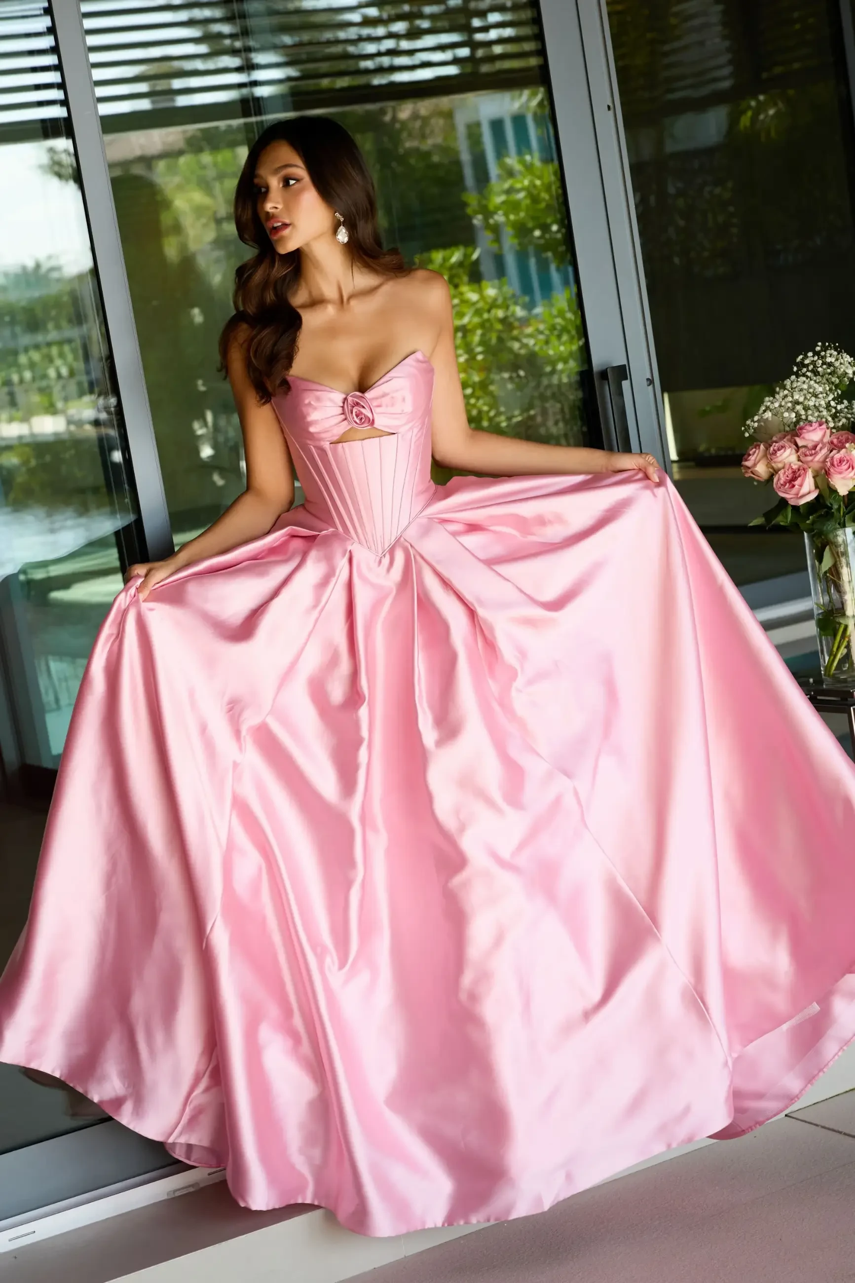A woman in an elegant, strapless pink gown stands in a modern interior. She holds the skirt wide, exuding grace. A bouquet of pink roses is nearby.