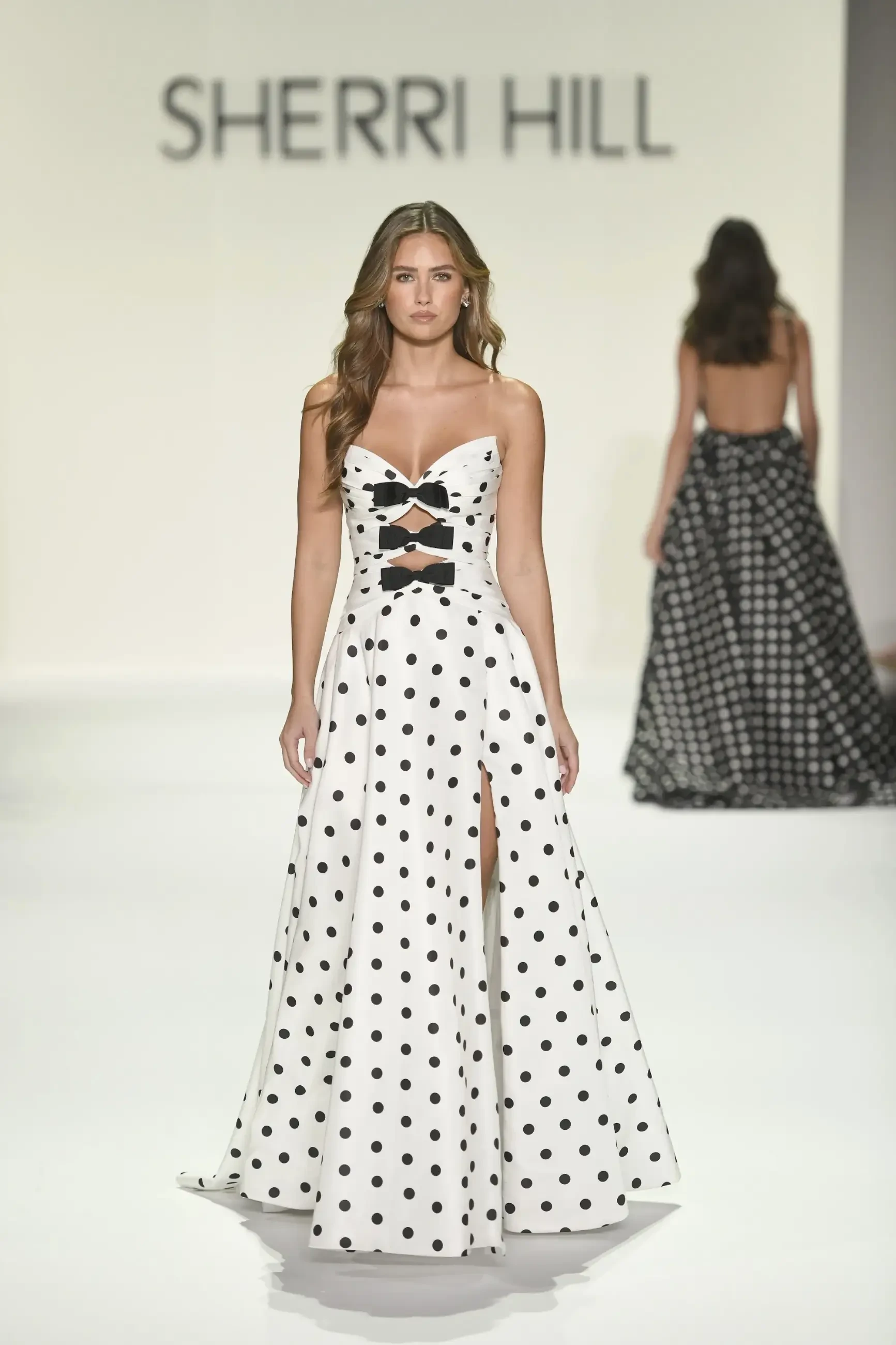 Model on runway wearing a white polka dot gown with cutouts and high slit. Background shows 