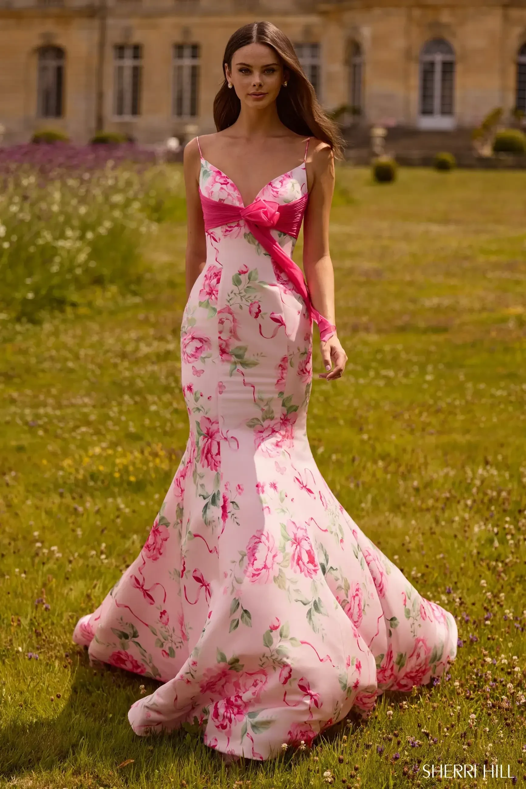 A woman is wearing a floral pink and white gown while standing in a grassy area.