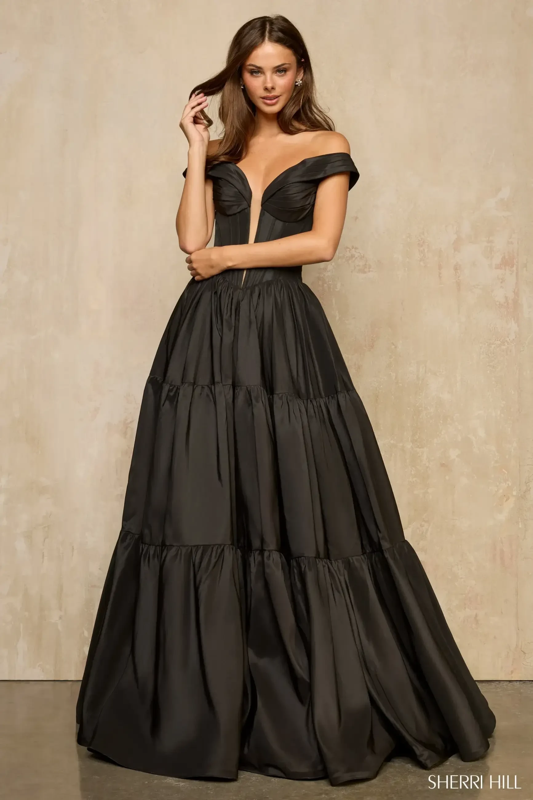 A model is wearing a black off-the-shoulder gown with a fitted bodice and a tiered skirt. The gown has a deep V-neckline and is floor-length, creating an elegant silhouette. The background is neutral, emphasizing the dress.