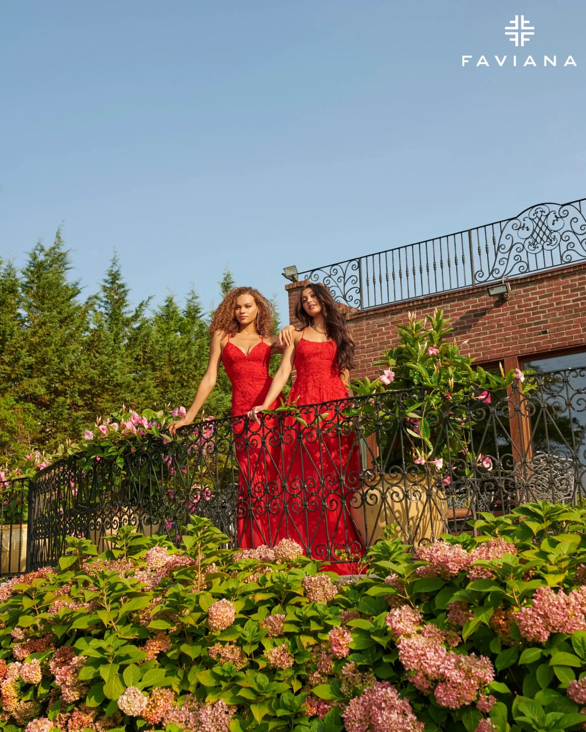 Two women in red dresses pose on a balcony surrounded by blooming flowers and greenery.